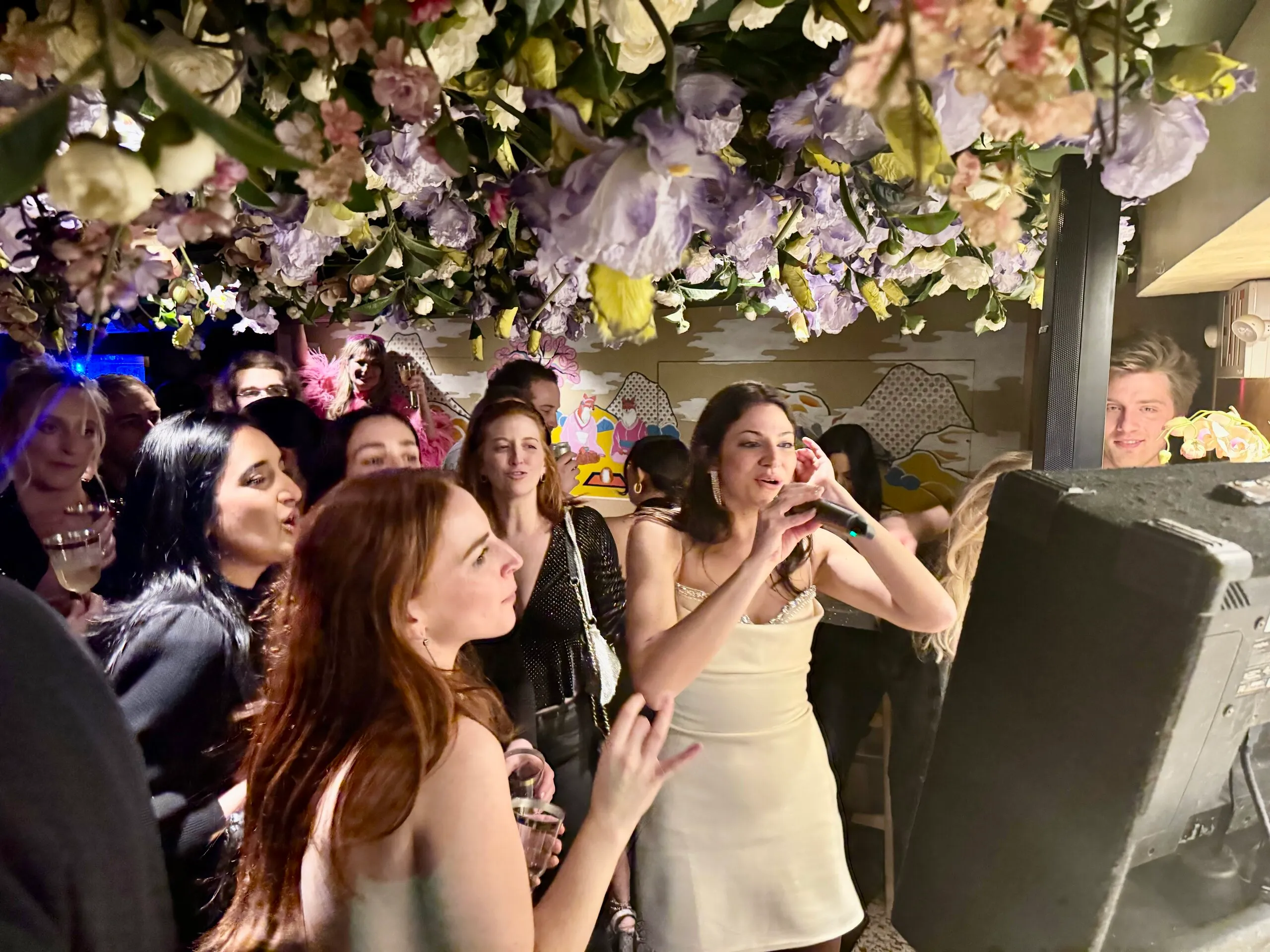 A young crowd singing karaoke together under a flower installation at a stylish venue