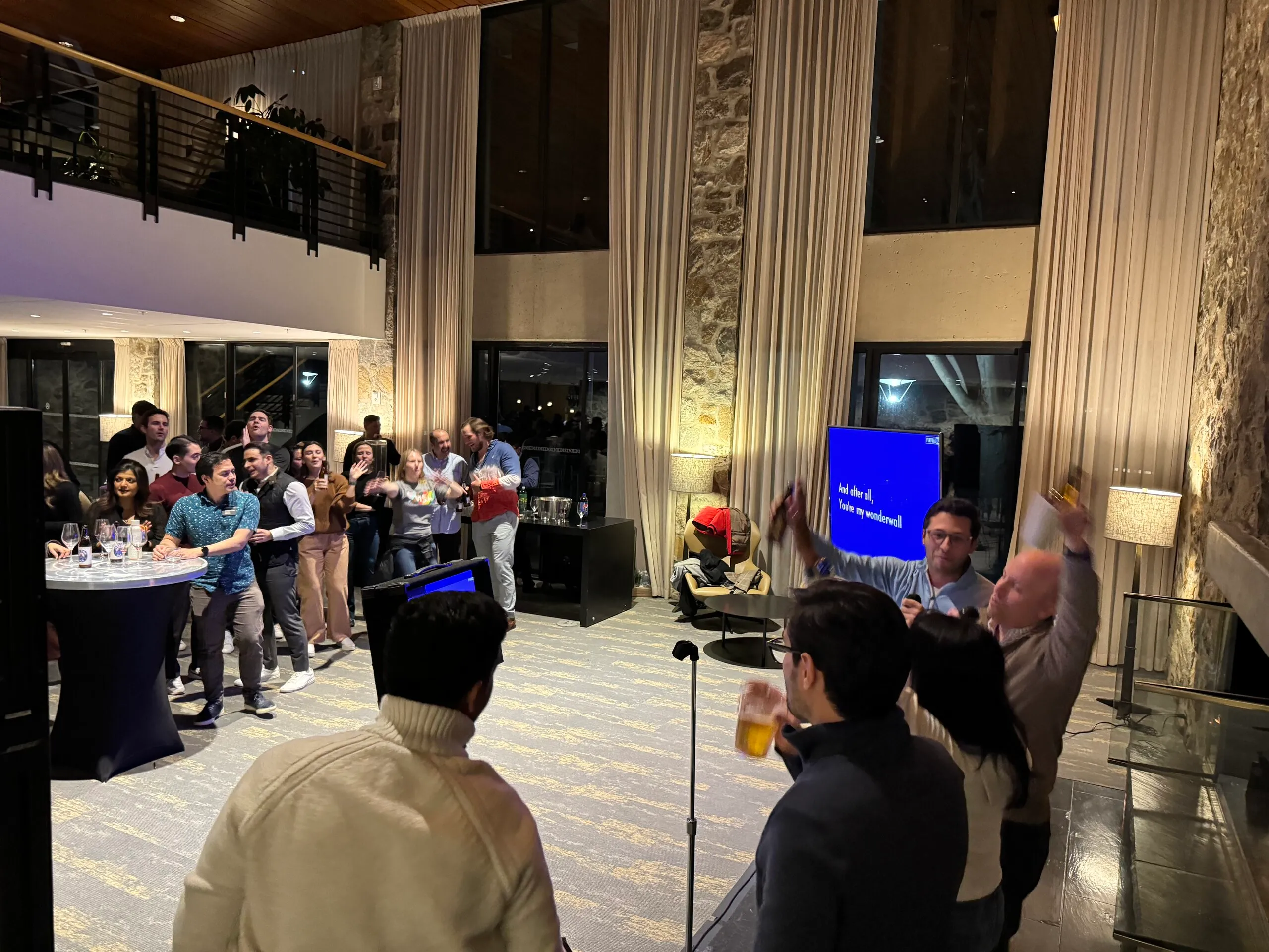 Guests singing karaoke together at an upscale corporate event, lyrics on the screen and drinks in hand