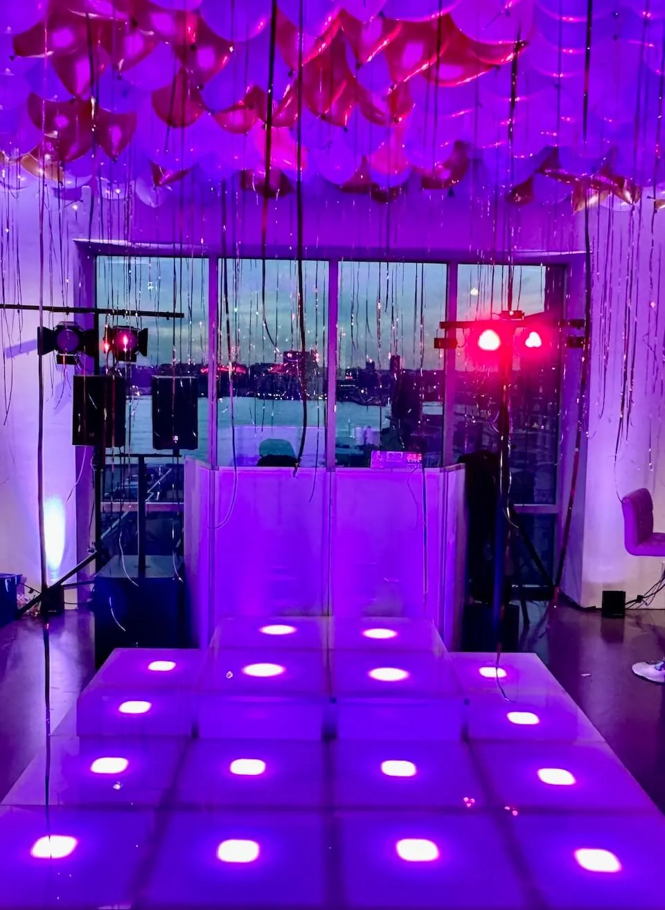 LED stage panels glowing purple and pink with uplighting and a DJ booth beside a waterfront window at dusk