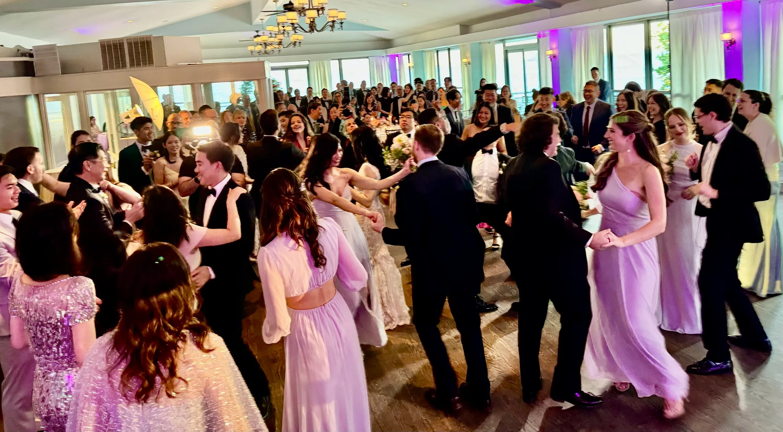 Guests in tuxedos and gowns dancing together on a packed wedding dance floor at an elegant waterfront ballroom